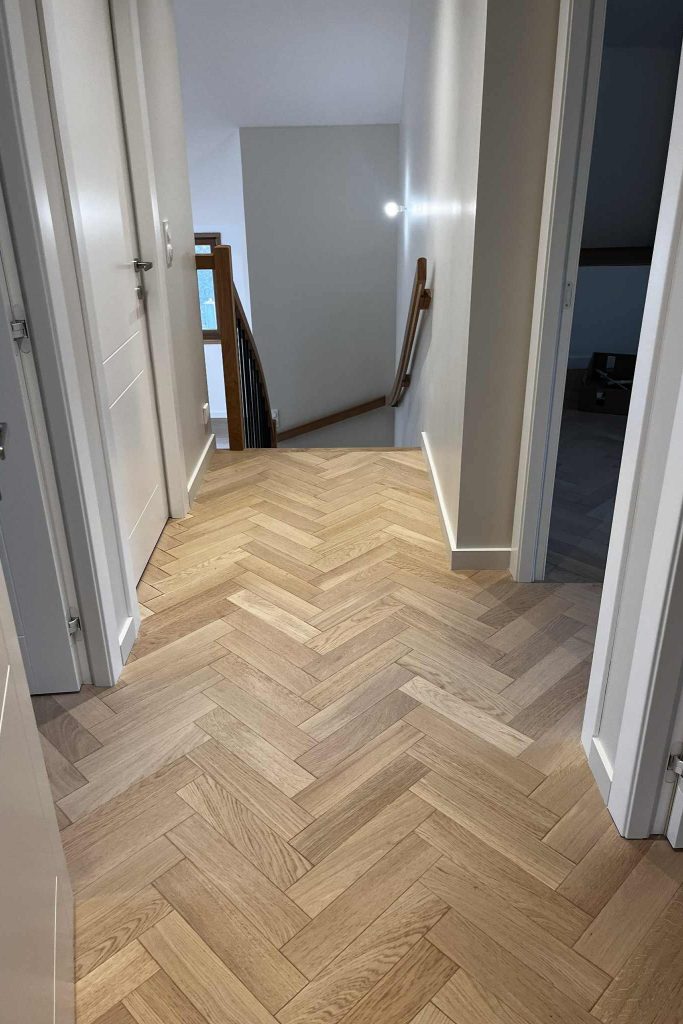 Un couloir moderne avec un sol en parquet à chevrons, des murs blancs et une rampe en bois, menant à un escalier.